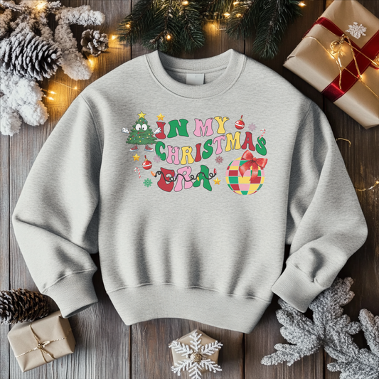 A light gray crewneck sweatshirt featuring the colorful "In My Christmas Era" design with festive icons like ornaments, a Christmas tree, and a present, surrounded by holiday decorations on a wooden background.