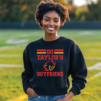 Model wearing a black 'Go Taylor's Boyfriend' sweatshirt featuring bold text and football heart design, standing on a football field.