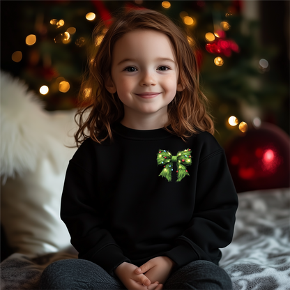 A smiling child wearing a black crewneck sweatshirt with a vibrant green bow design, inspired by the Grinch, sitting in front of a decorated Christmas tree.