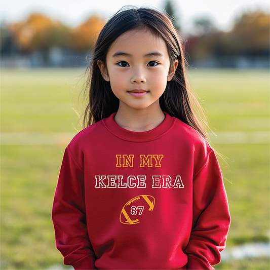 Child wearing a red 'In My Kelce Era' sweatshirt with football graphic, standing on a grassy field.