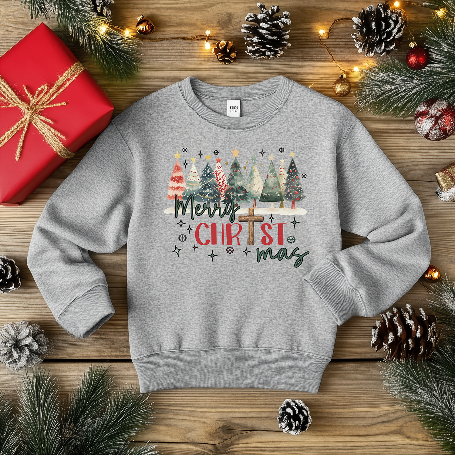 A light gray crewneck sweatshirt with the "Merry CHRISTmas" design, showcasing festive Christmas trees and a cross, styled with pinecones, Christmas lights, and gift decor.