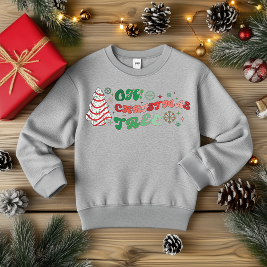 A light gray crewneck sweatshirt featuring the "Oh Christmas Tree" design with a decorated tree, colorful snowflakes, and festive text, styled with holiday decorations, pinecones, and Christmas lights.