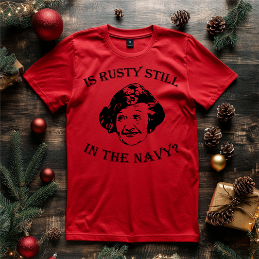 "Is Rusty Still in the Navy?" holiday shirt featuring a humorous design inspired by a classic movie moment, styled on a red T-shirt surrounded by festive pinecones and decorations.