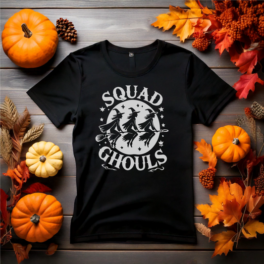 Black Halloween-themed t-shirt with 'Squad Ghouls' design featuring three witches flying on brooms, surrounded by stars. Laid flat with autumnal decorations, including pumpkins and fall leaves, capturing a spooky seasonal vibe. Perfect for group Halloween outfits or fun fall gatherings.