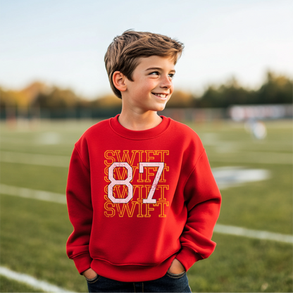 Child wearing a red '87 Swiftie' sweatshirt featuring bold text and football design, standing on a sports field.