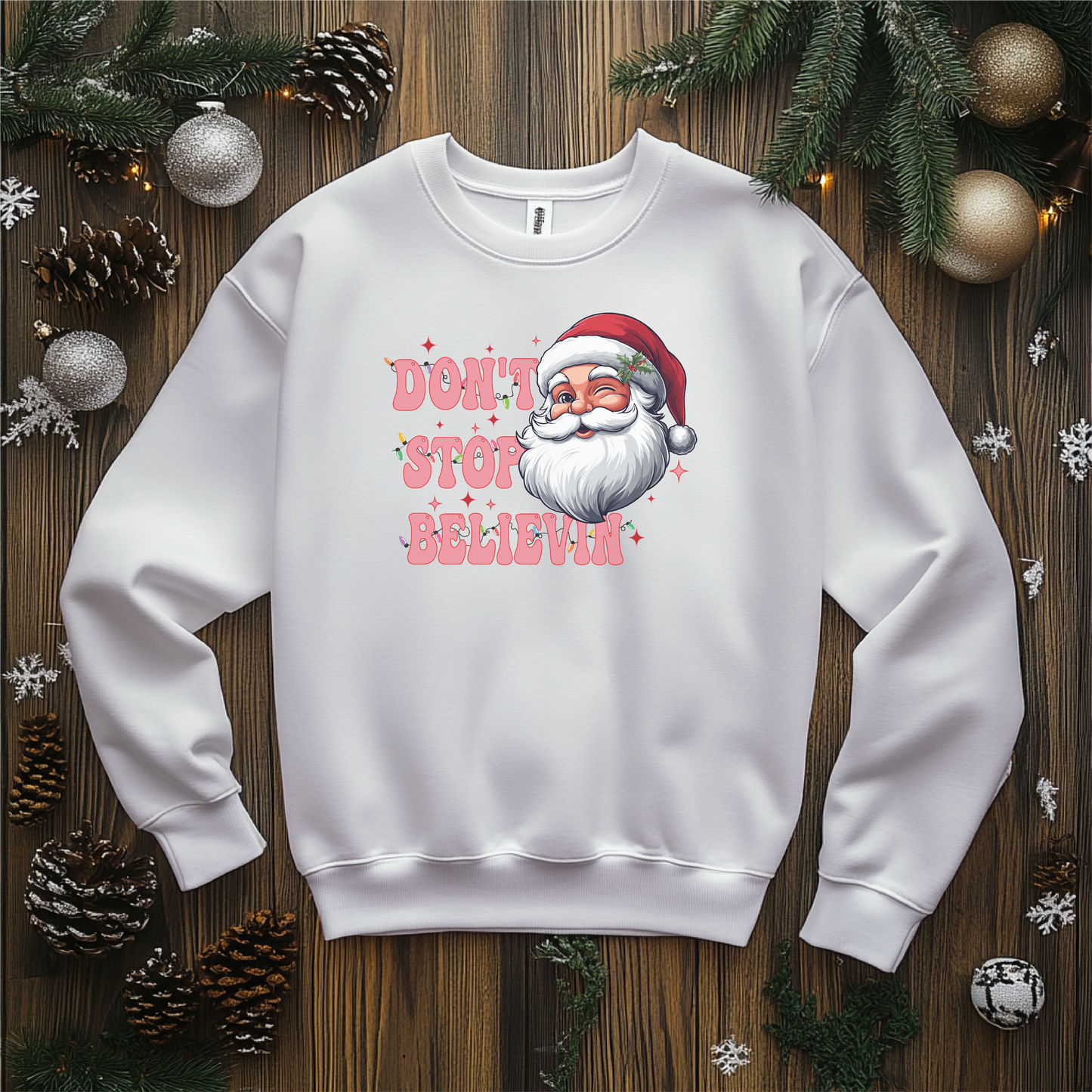 Don't Stop Believin" Christmas shirt featuring a retro Santa design with playful typography, styled on a white crewneck surrounded by festive pinecones, ornaments, and greenery.